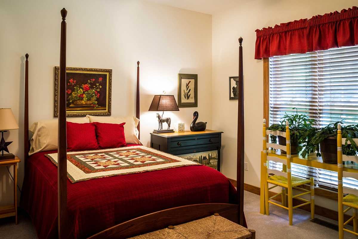 Chambre meublée avec lit à colonnes en bois, couvre-lit rouge et mobilier traditionnel, fenêtre avec rideau rouge Chambre meublée avec lit à colonnes en bois, couvre-lit rouge et mobilier traditionnel, fenêtre avec rideau rouge