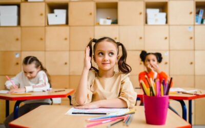 Une élève lève la main au bureau, deux camarades écrivent, casiers derrière