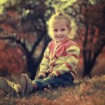 Une petite fille assise sur l'herbe, souriante, en pull rayé, avec des arbres automnaux
