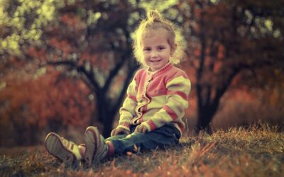 Une petite fille assise sur l'herbe, souriante, en pull rayé, avec des arbres automnaux