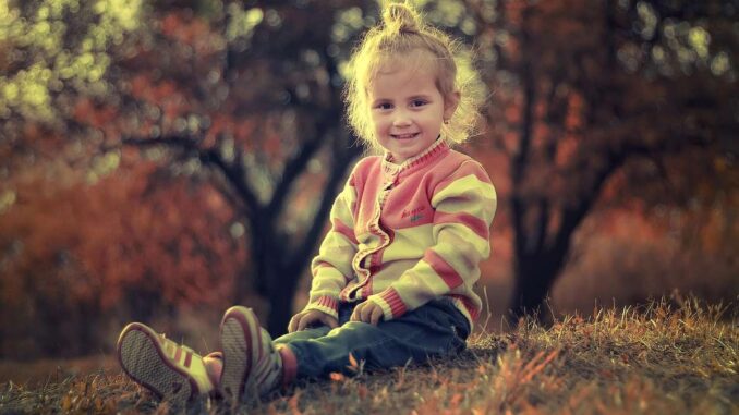 Une petite fille assise sur l'herbe, souriante, en pull rayé, avec des arbres automnaux